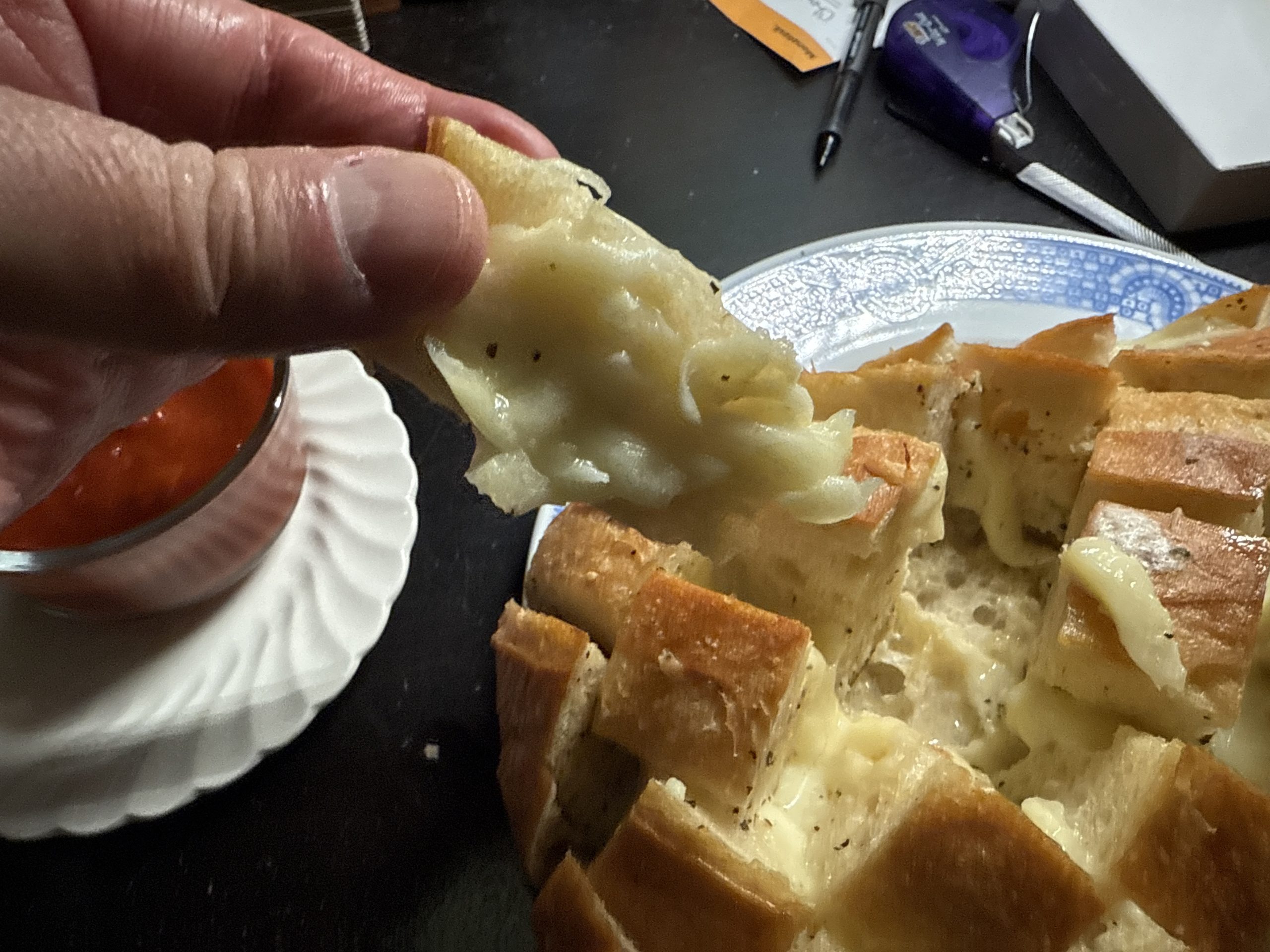 A hand appears from outside the left frame reaches and tears a chunk of bread from a cheese and garlic loaf on a blue and white plate. Nearby sits a white ceramic dish of marinara for dipping. The setting for the meal as explained in the accompanying post is a personal gratitude celebration.