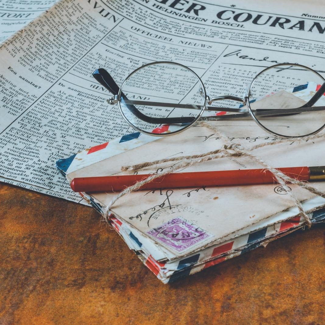 Historical letters and a fountain pen on a vintage wooden table, alongside eyeglasses and a newspaper.